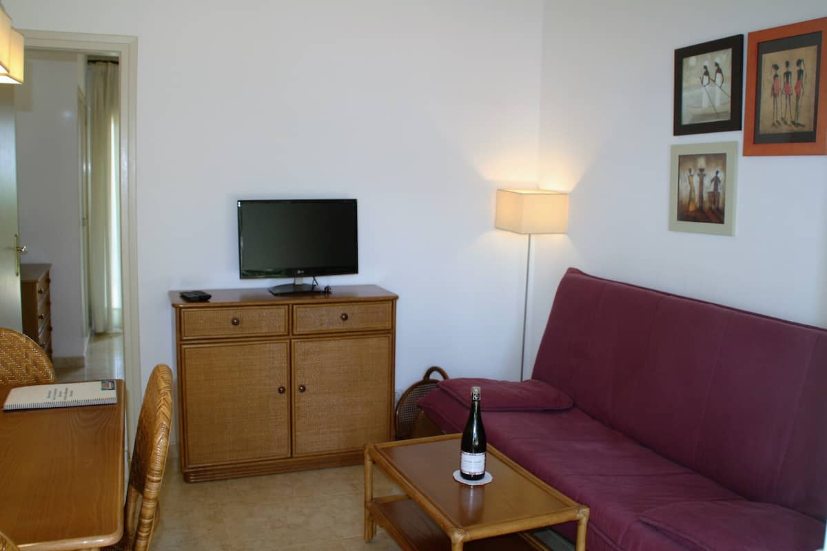 Una sala de estar con televisor sobre un mueble de madera, un sofá rojo, una mesa de centro de madera con una botella de vino y una lámpara.