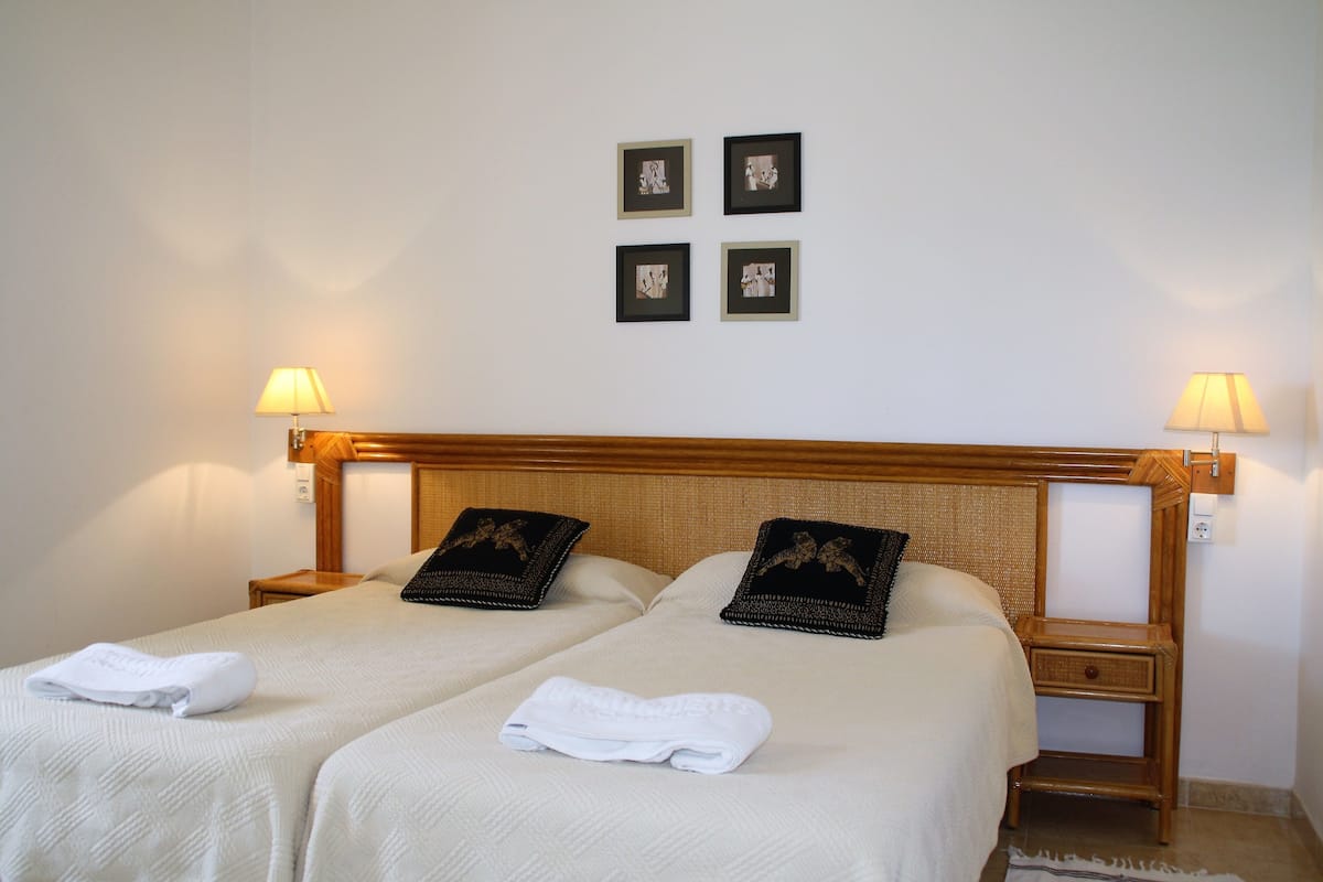 Habitación de hotel con dos camas, cada una con una almohada y una toalla doblada. Hay dos mesitas de noche con lámparas y una pequeña mesita auxiliar de madera.