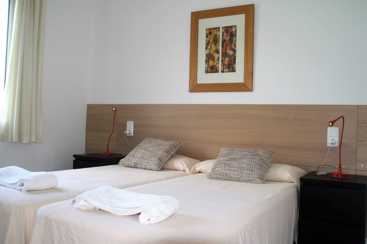 Habitación de hotel con dos camas, cabecero de madera, una obra de arte enmarcada y lámparas de noche.