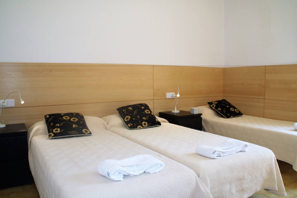 Habitación de hotel con dos camas individuales, cada una con una almohada y una toalla doblada. Hay mesitas de noche con lámparas y un enchufe en la pared.