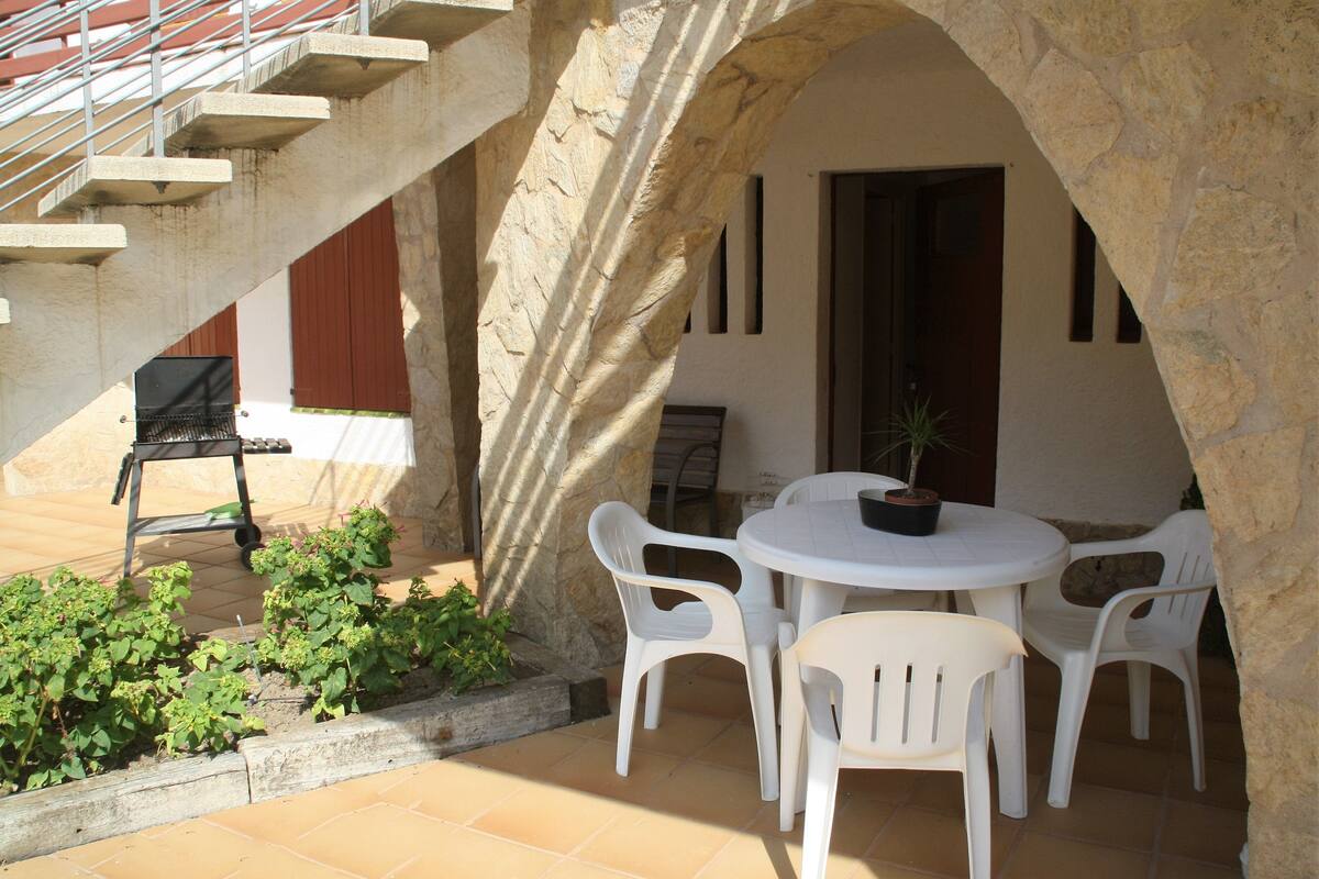 Un patio con una mesa redonda y sillas de plástico blancas, una planta en maceta y una escalera que lleva a un nivel superior.