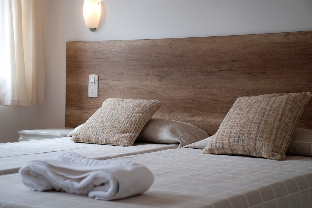 Una cama bien hecha con cabecera de madera, dos almohadas y una toalla blanca enrollada.