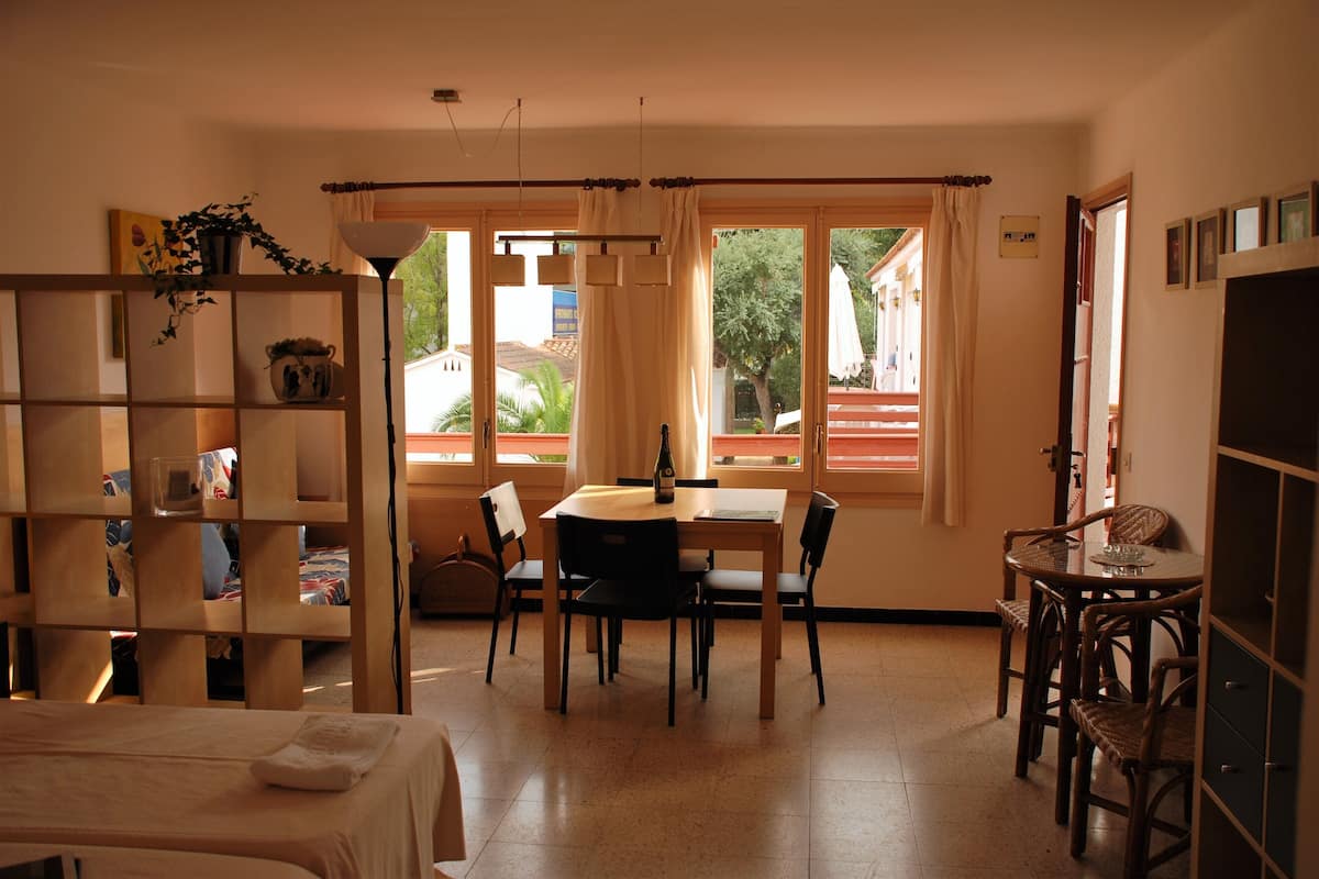 Una habitación con cama, una mesa de comedor con sillas, una estantería y ventanas amplias con cortinas.
