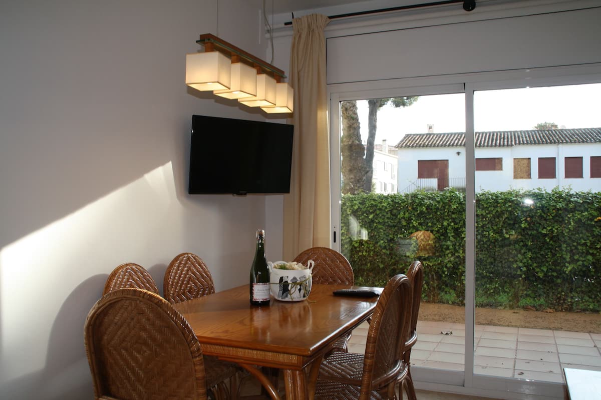 Un comedor con una mesa de madera, sillas de mimbre, una botella de vino y un cuenco de fruta. Un televisor de pantalla plana está montado en la pared sobre la mesa.