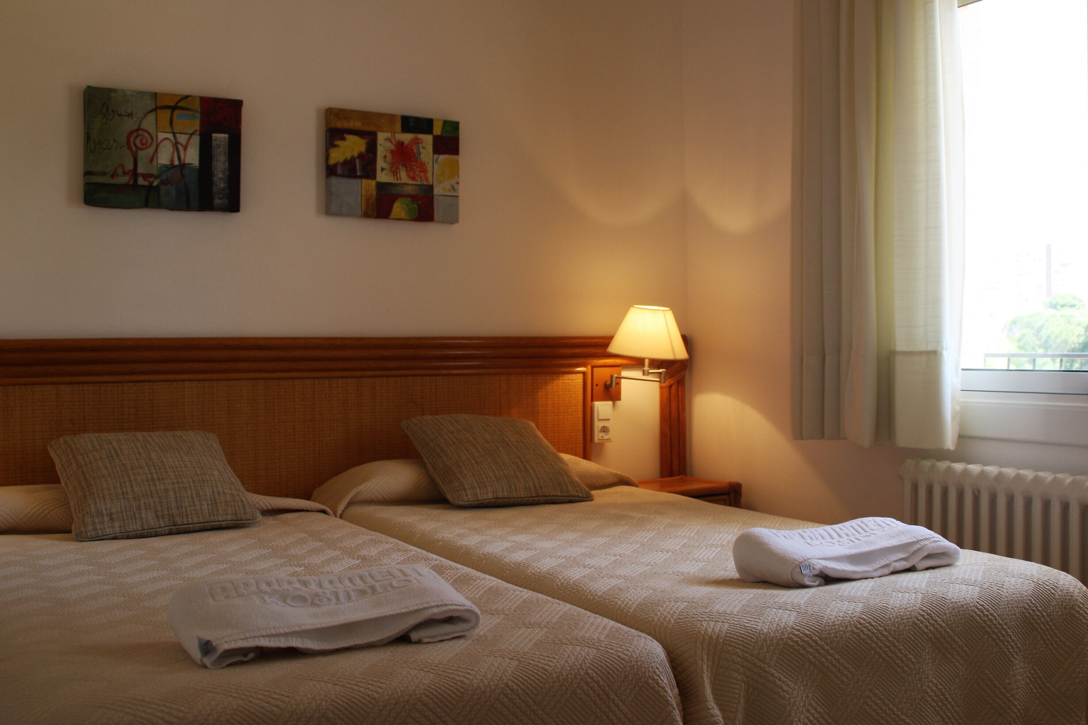A hotel room with two beds, a nightstand with a lamp, and two paintings on the wall.
