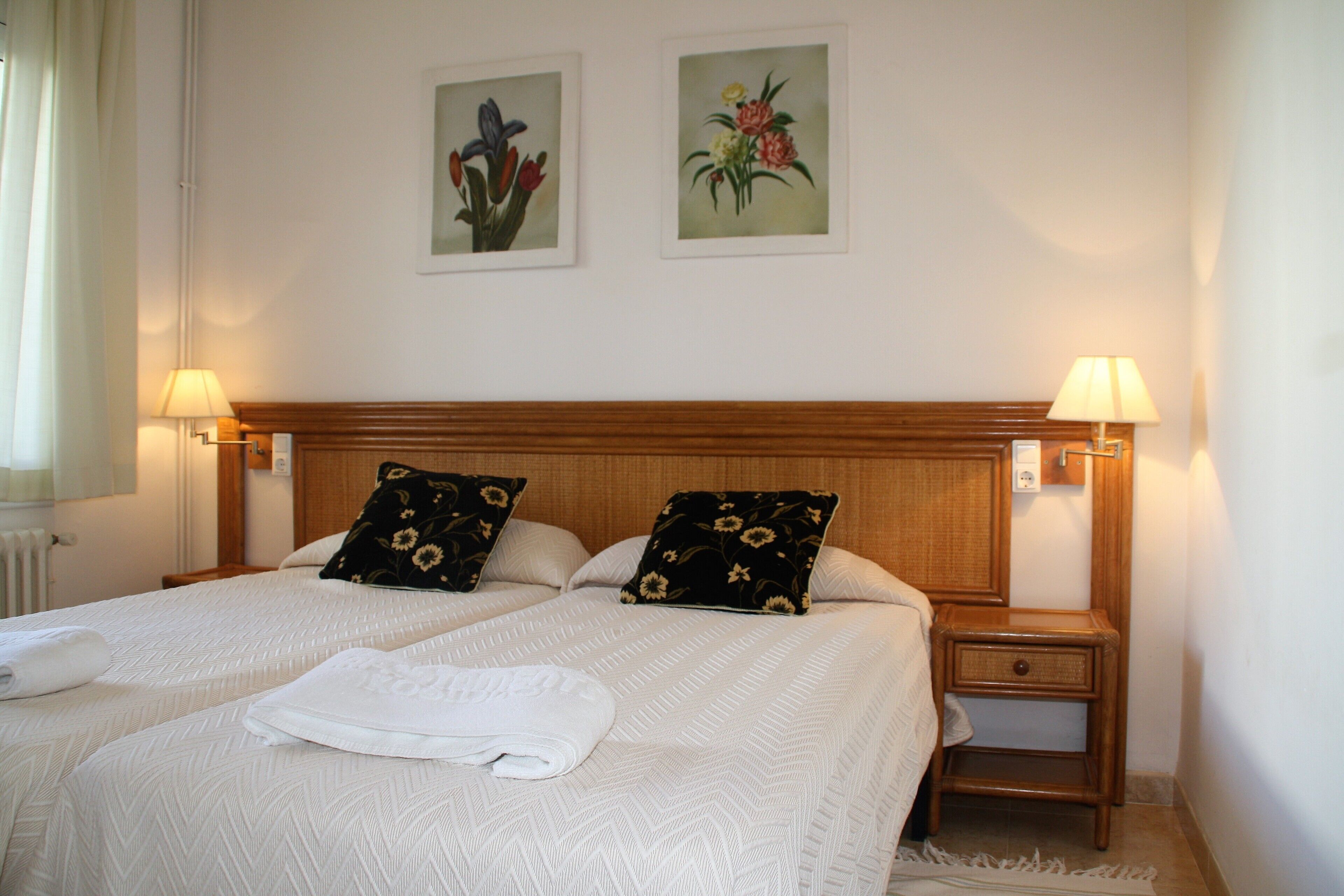 A double bed with a white bedspread and two black pillows with floral patterns. There are two wall-mounted lamps on either side of the bed.