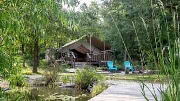 A rustic cabin with a wooden deck, surrounded by lush greenery and a small pond.