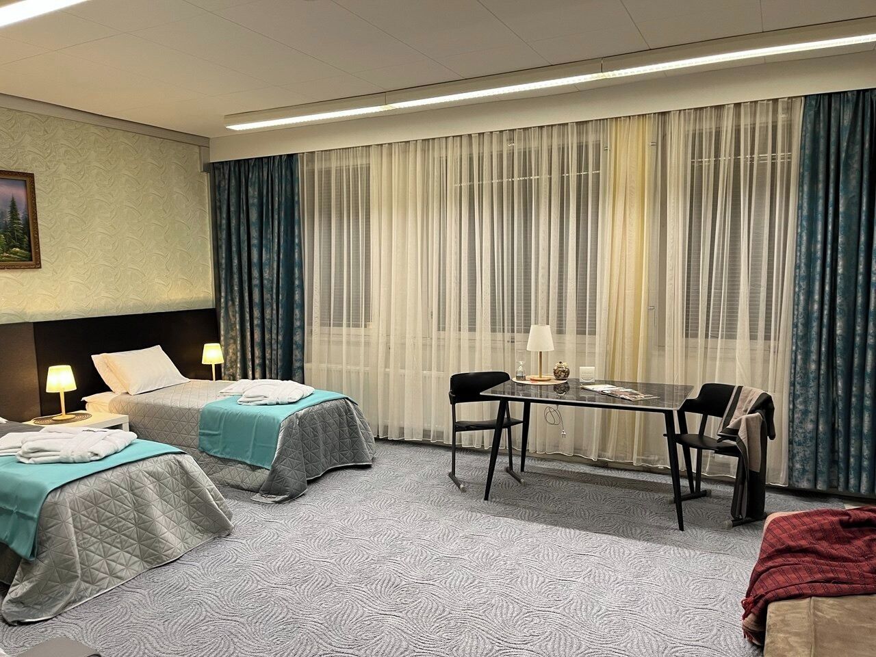 Habitació d'hotel amb dos llits, una taula petita, cadires, una làmpada i una finestra gran amb cortines.