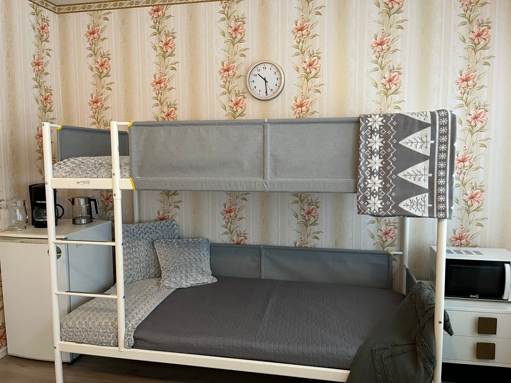 Un lit superposé avec un matelas gris et des oreillers à motifs, une petite table avec une bouilloire et une machine à café, et un four à micro-ondes posé sur un meuble.