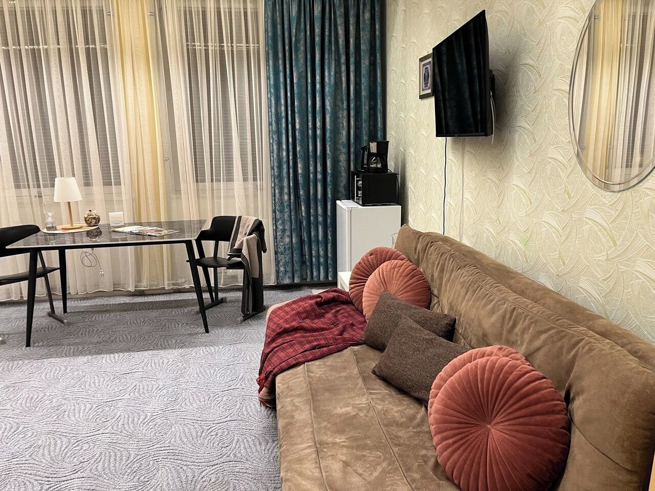 A living room with a brown sofa, red and brown cushions, a coffee table, a TV mounted on the wall, and a patterned carpet.