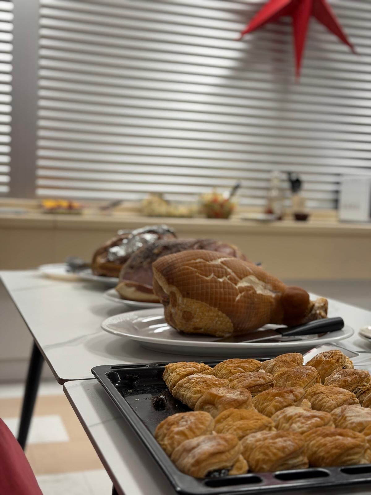 Una mesa con pasteles y jamón en una bandeja, con una ventana y persianas en el fondo.