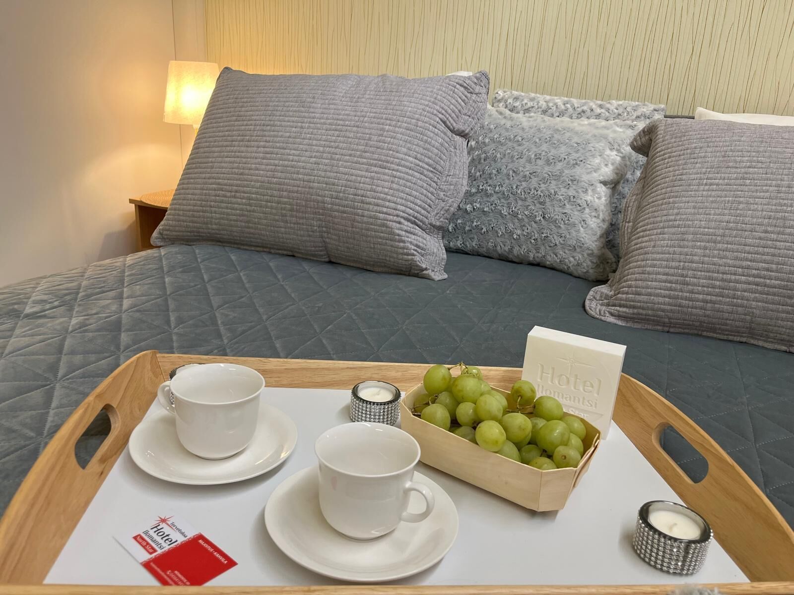 Una bandeja de desayuno ordenada con dos tazas, una caja de uvas verdes y una vela sobre una cama con ropa de cama y almohadas grises.