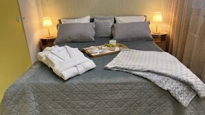 Una cama bien hecha con colcha gris, dos almohadas, una bandeja con una taza, un bol de fruta y toallas dobladas.