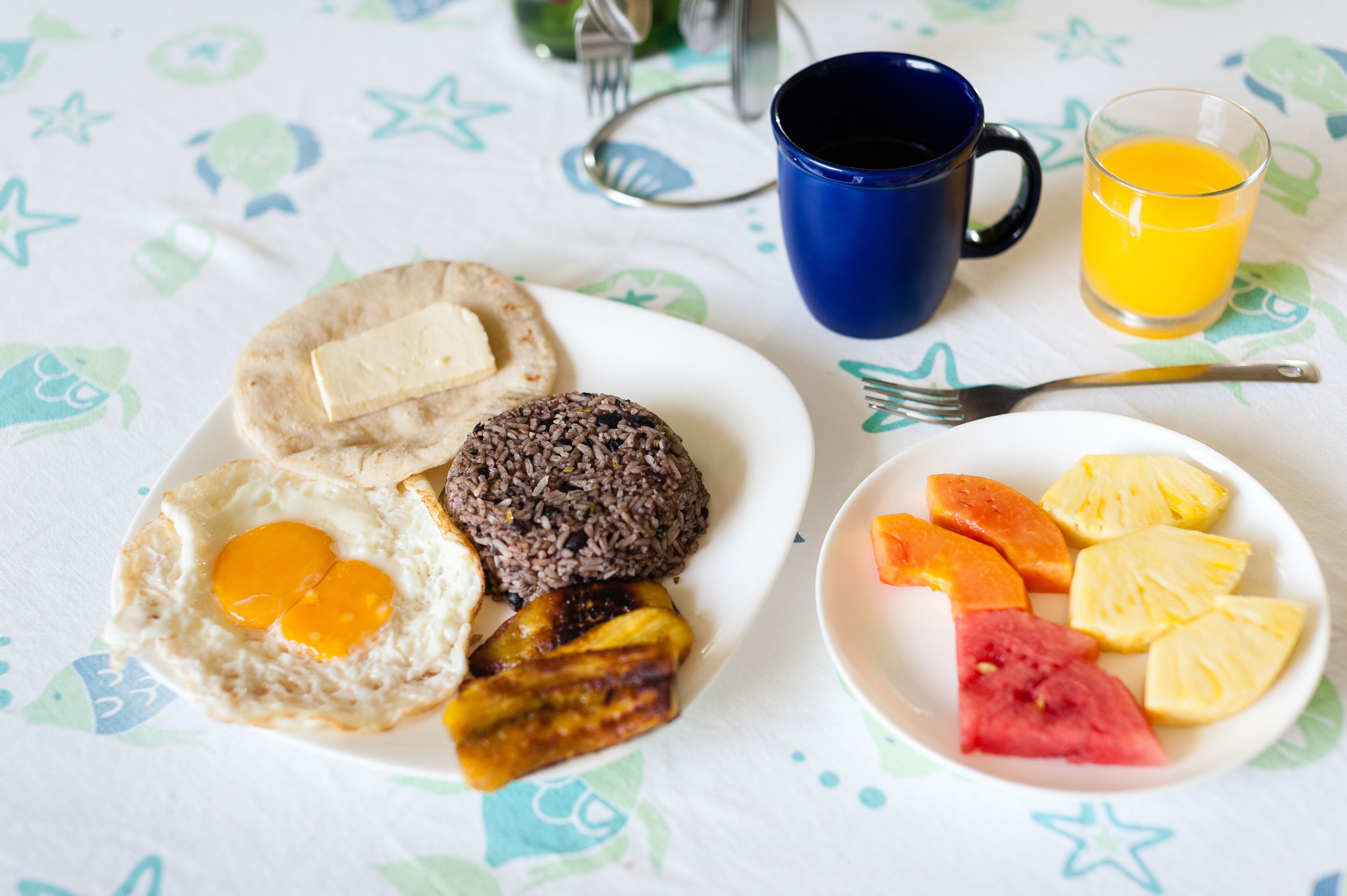 Un plato de desayuno con huevos, tostadas, morcilla y plátanos fritos, un bol de fruta y un vaso de zumo de naranja sobre una mesa con mantel estampado de estrellas.