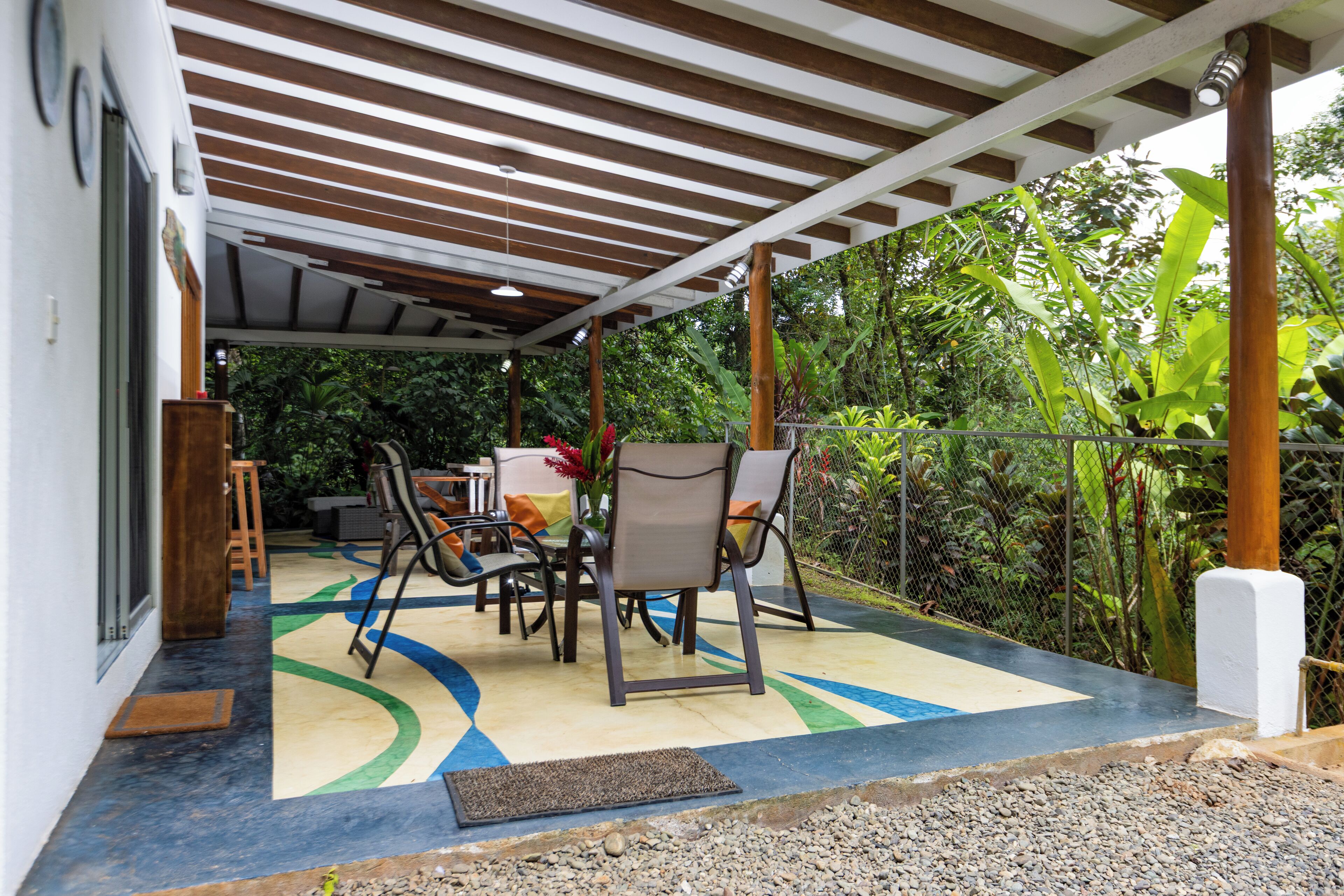 Un patio exterior con una mesa y sillas, rodeado de vegetación y una zona cubierta con vigas de madera.