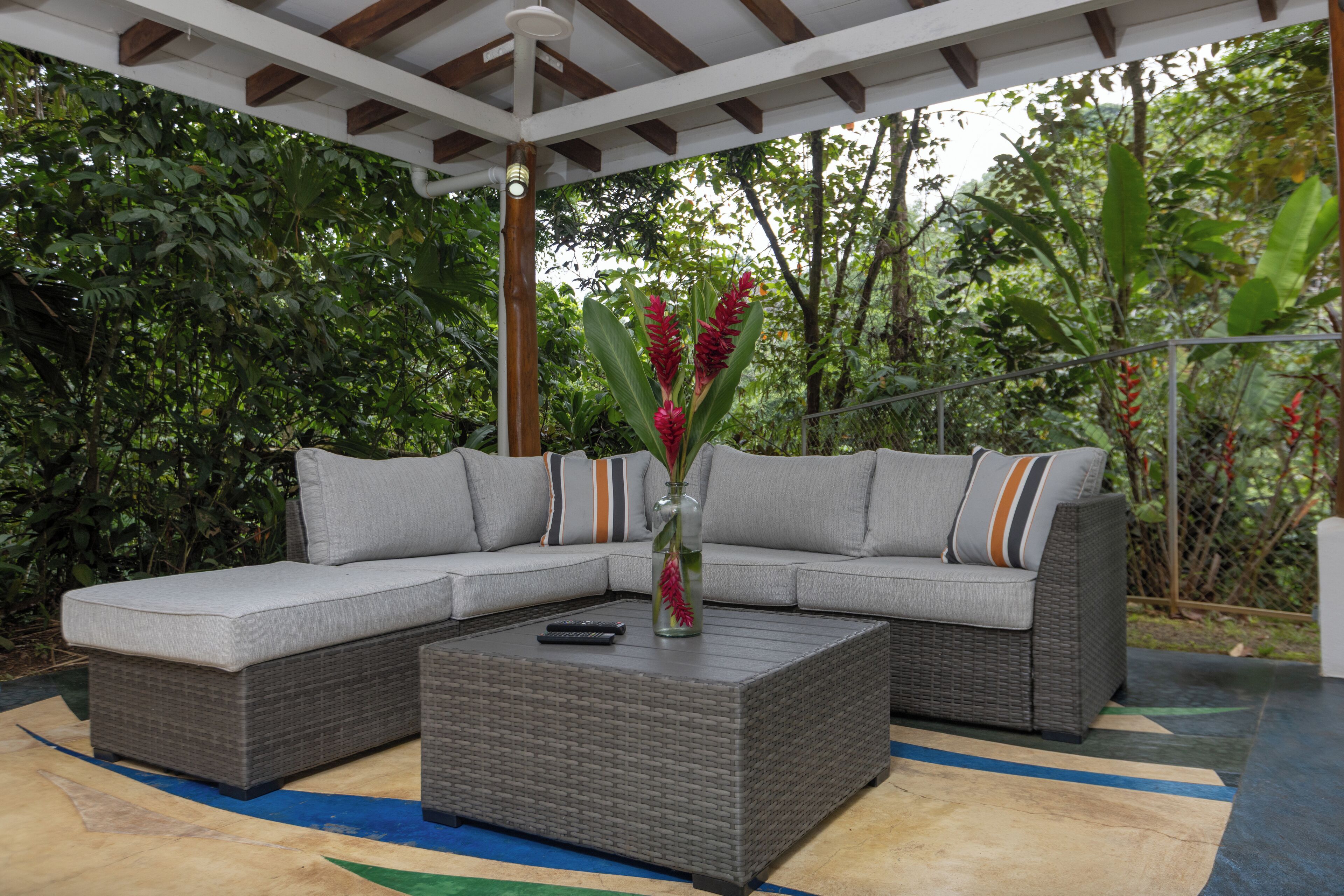 A covered outdoor seating area with a grey sectional sofa, a woven coffee table, and a vase with red flowers.