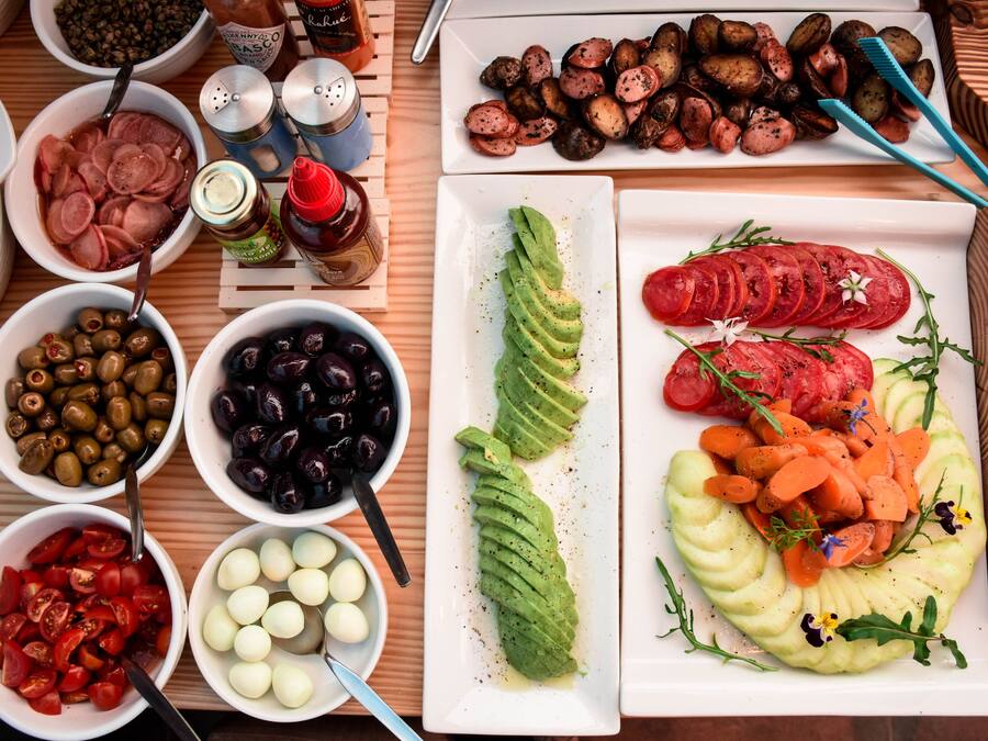 Diversitat d'aliments com ara talls d'alvocat, tomàquets, olives i ous disposats sobre una taula de fusta.
