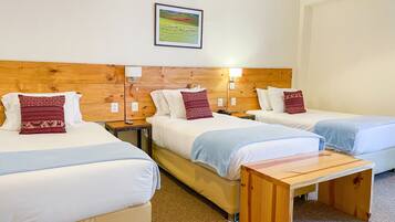 Habitación de hotel con tres camas, cada una con cabecero de madera, ropa de cama blanca y almohadas estampadas. Entre las camas hay una mesita de noche de madera con una lámpara, y una pequeña mesa de centro.