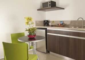 Een compacte keuken met een magnetron, koffiezetapparaat en een klein spoelbakje. Er staat een eettafel met stoelen en een vaas met bloemen.