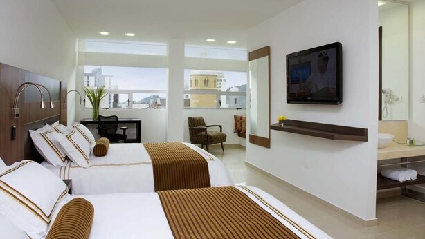 A hotel room with two beds, a desk, a chair, a TV, and a bathroom with a mirror and sink. - San Simon Hotel Boutique (Pereira)