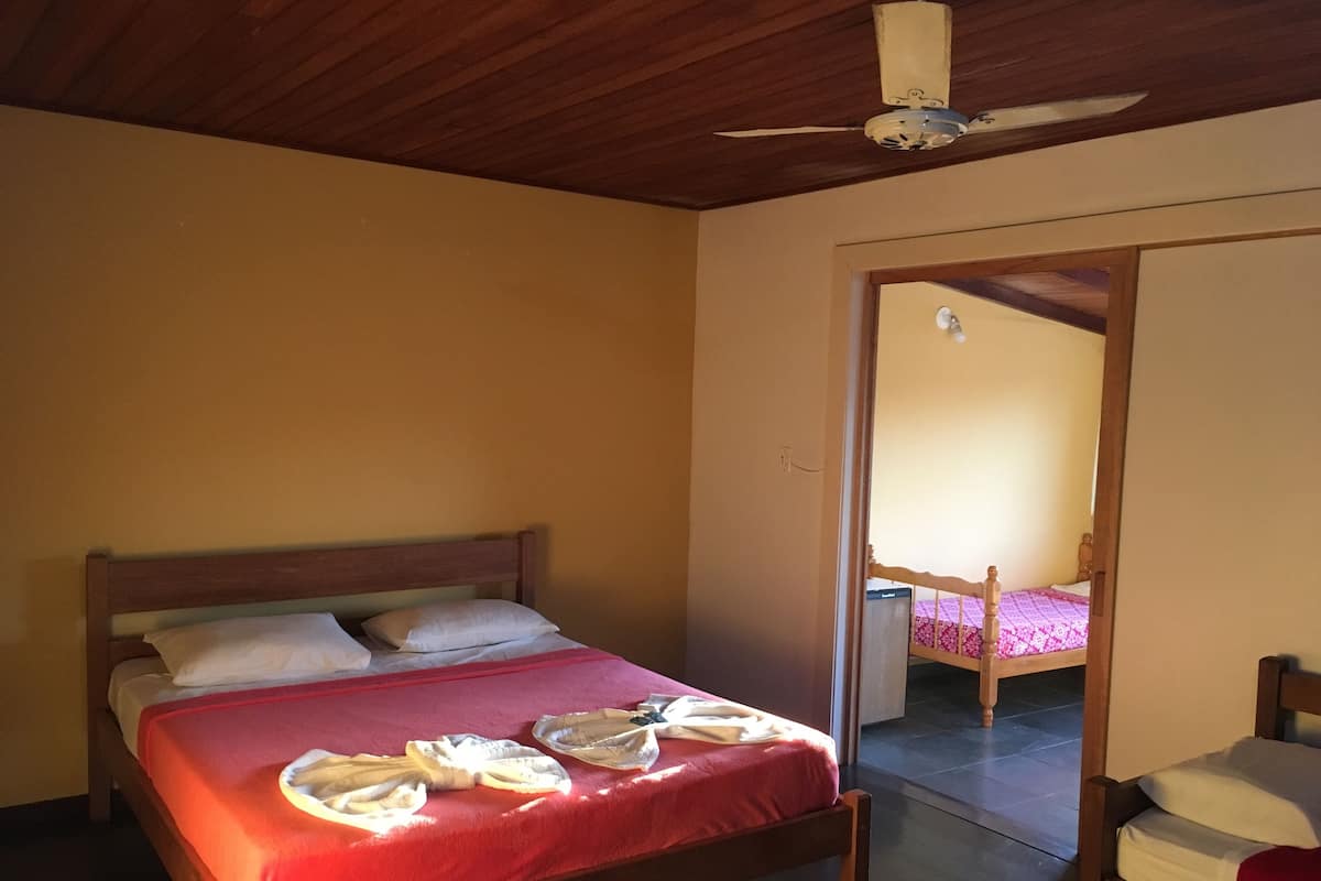 Un lit simple avec une couvre-lit rouge et des oreillers blancs, un ventilateur de plafond et une tête de lit en bois.