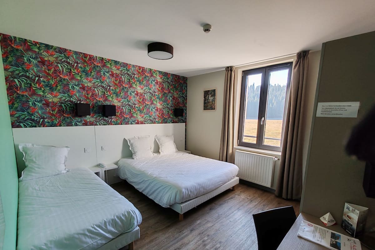 Habitación de hotel con dos camas, papel tapiz floral, ventana con vistas y mesita de noche con lámpara.