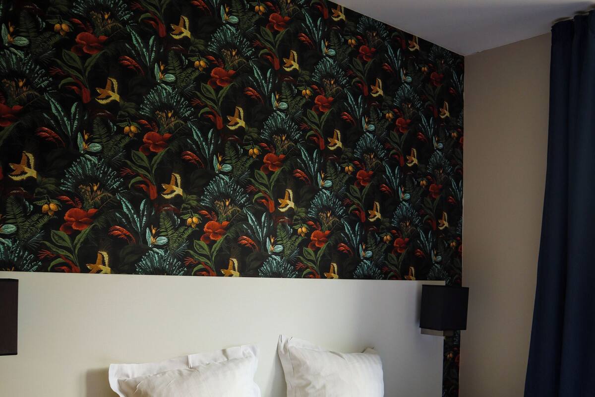 Habitación de hotel con cama, dos almohadas blancas, mesita de noche, lámpara y papel tapiz estampado con flores.