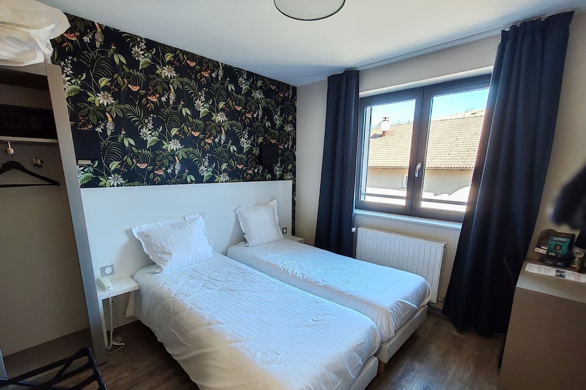 Una habitación con dos camas, un radiador, una ventana con cortinas, una pared con diseño floral y una mesita con lámpara.