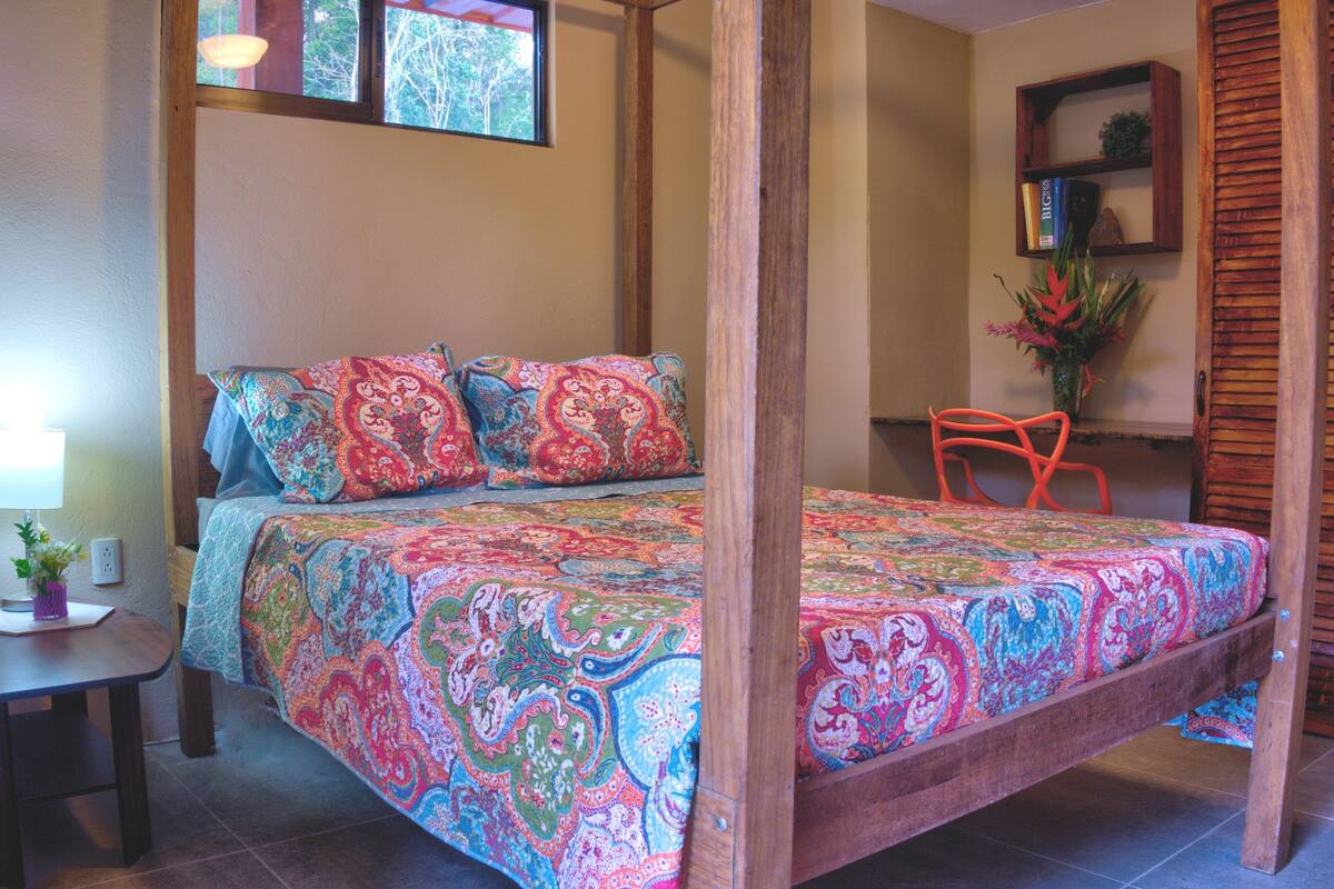 A four-poster bed with a colorful, patterned bedspread, a wooden chair, a small table with a lamp, and a window with a view of trees.