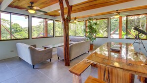 A spacious room with a wooden ceiling, large windows, a sofa, a coffee table, and a wooden dining table. - Valley of Dreams Eco Lodge Room (Barú)