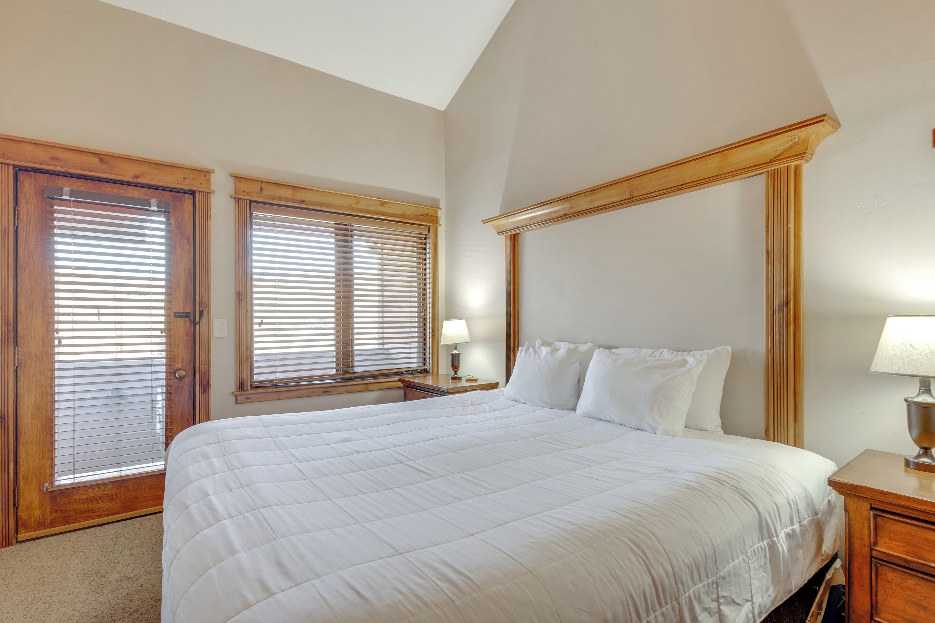 A bedroom with a large bed, wooden headboard, two windows with blinds, a nightstand with a lamp, and a carpeted floor.