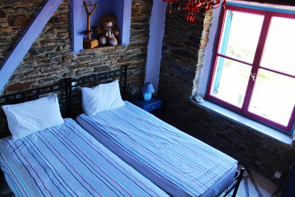 A bedroom with a stone wall, a bed with striped bedding, a nightstand with a lamp, and a window with a view.
