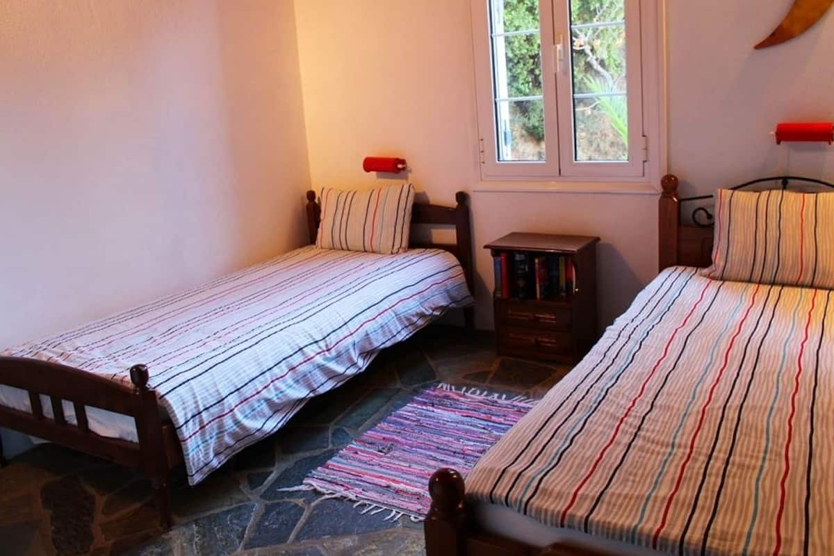 A bedroom with two single beds, a small wooden nightstand, a window with a view of greenery, and a stone floor.