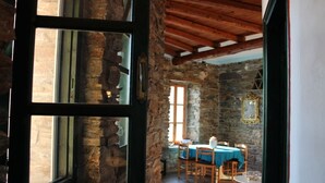 A rustic stone interior with a wooden ceiling, a dining area with a table and chairs, and a window with a view of the outside. - Villette Adonis (Andros)