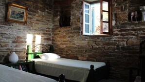 A rustic room with stone walls, a bed with a green headboard, a bedside table with a lamp, and a window with an open shutter. - Villette Adonis (Andros)
