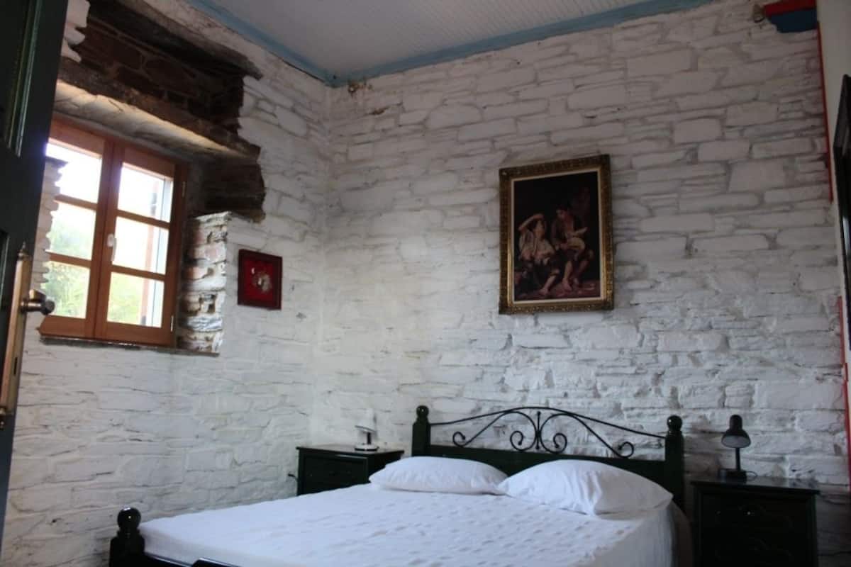 A bedroom with a stone wall, a bed with white bedding, a nightstand, a lamp, and a window with wooden shutters.