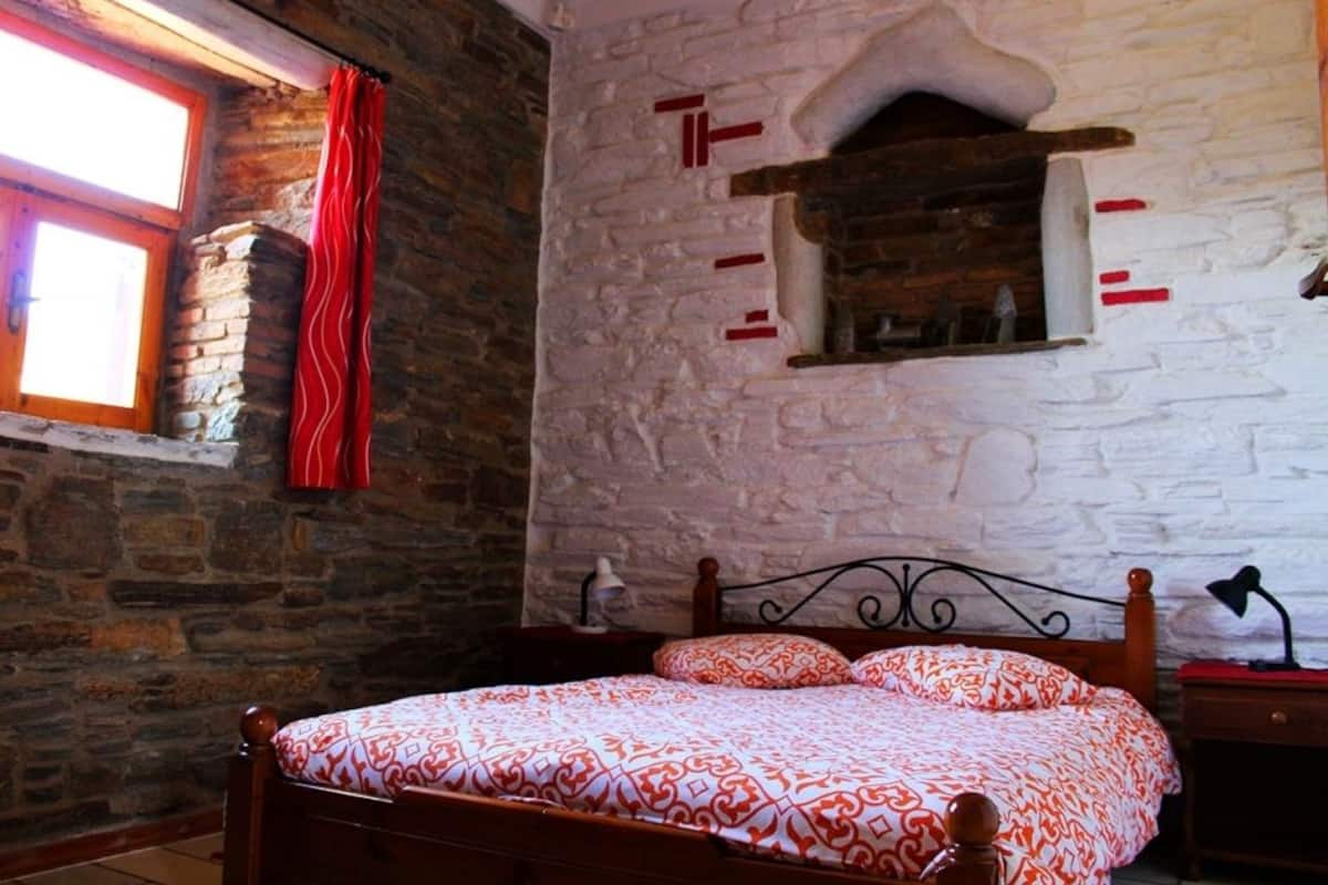 A bedroom with a stone wall, a wooden bed, a window with red curtains, and a small fireplace.