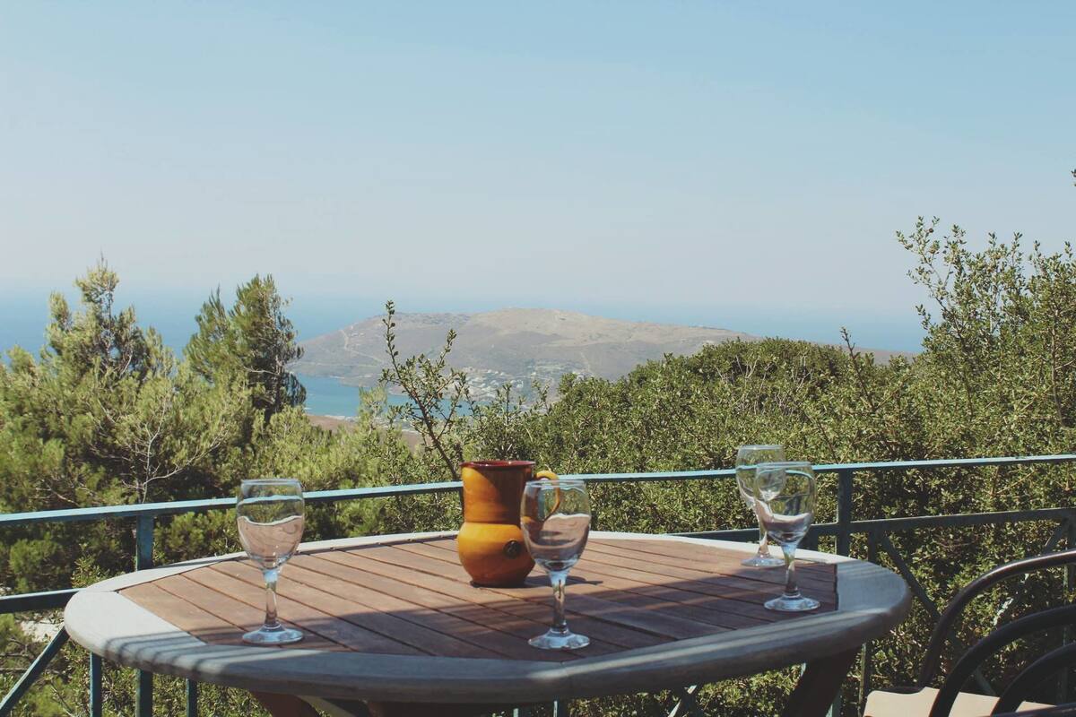 A wooden table with three wine glasses and a vase on a balcony overlooking a mountainous landscape.