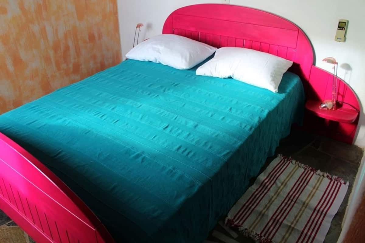 A bed with a teal bedspread and a red headboard. There is a bedside table with a lamp and a red chair.