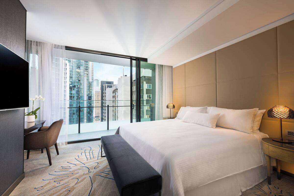 A modern hotel room with a large bed, a desk with a chair, a bench, a TV, and a view of the city through large windows.