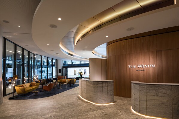 A modern hotel lobby with a curved reception desk, seating area with sofas and chairs, and large windows offering views of the city. - The Westin Brisbane (Brisbane)