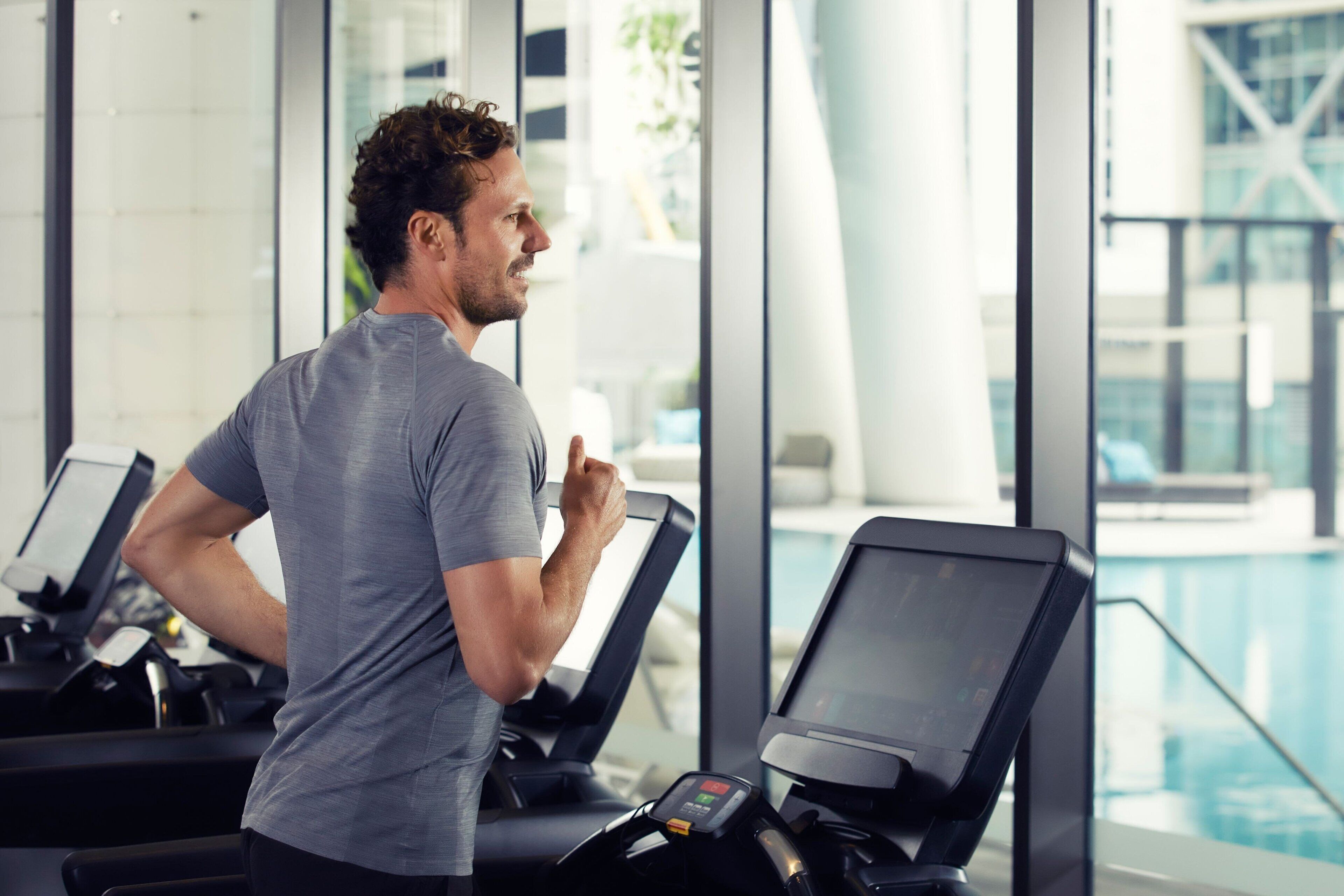A man using a treadmill in a gym with a pool visible through the windows.