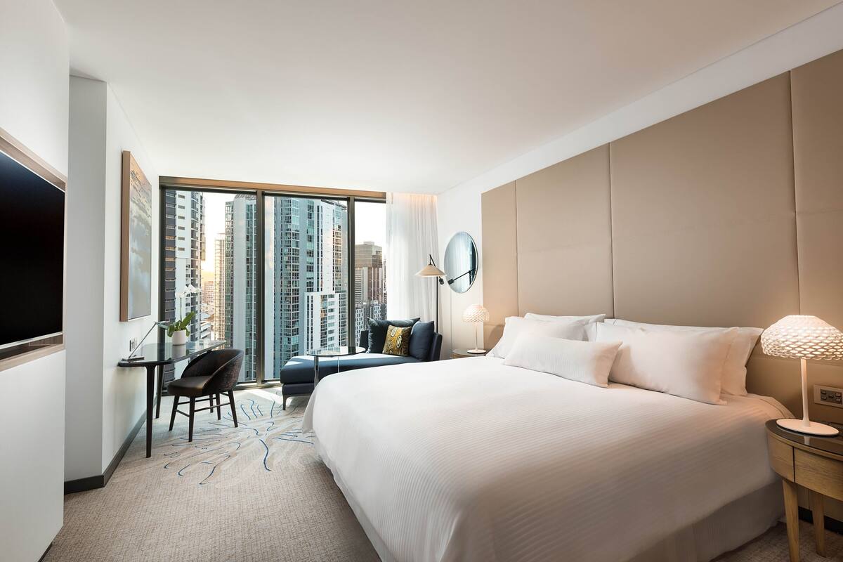 A modern hotel room with a large bed, a desk with a chair, a TV, and a view of tall buildings.
