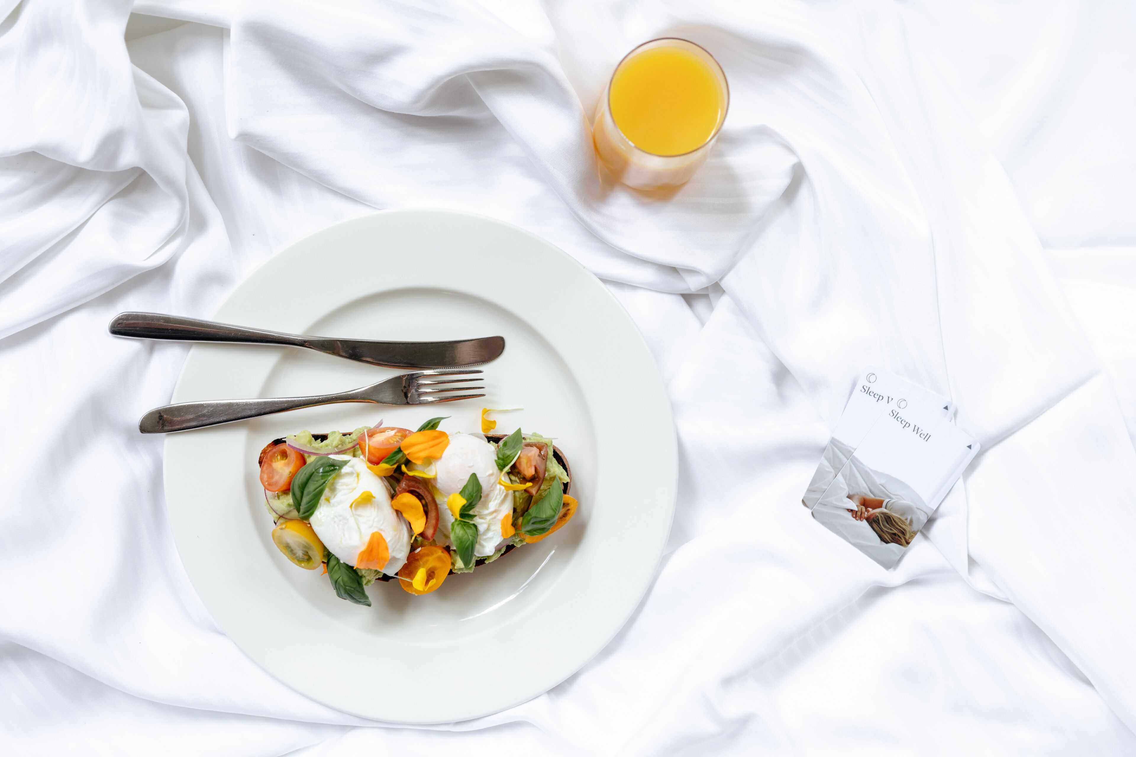 Un plato blanco con ensalada, un tenedor, un cuchillo y un vaso de jugo de naranja sobre un cubrelecho blanco.