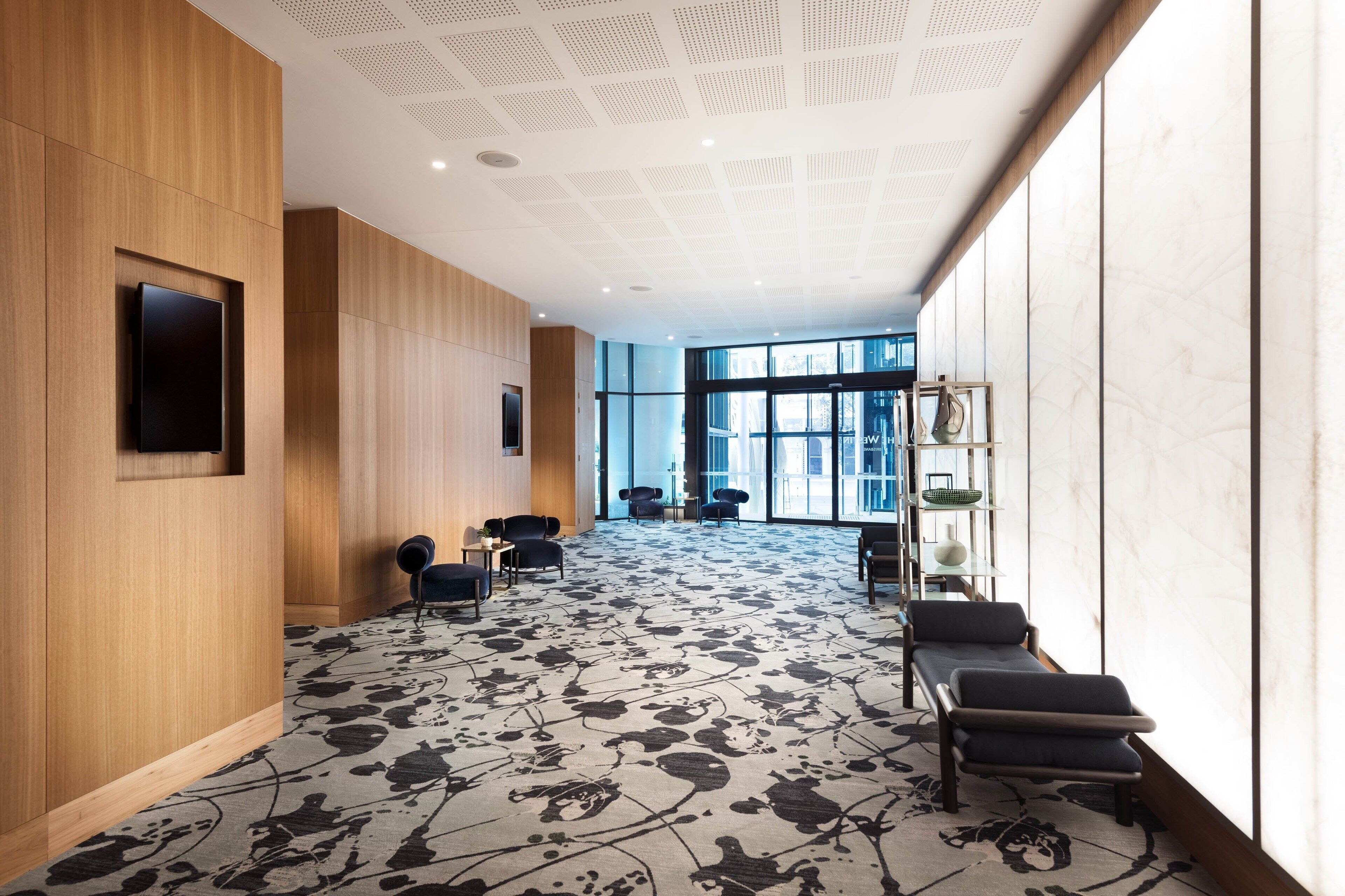 A modern hotel lobby with a patterned carpet, wooden panel walls, and a seating area with chairs and tables.