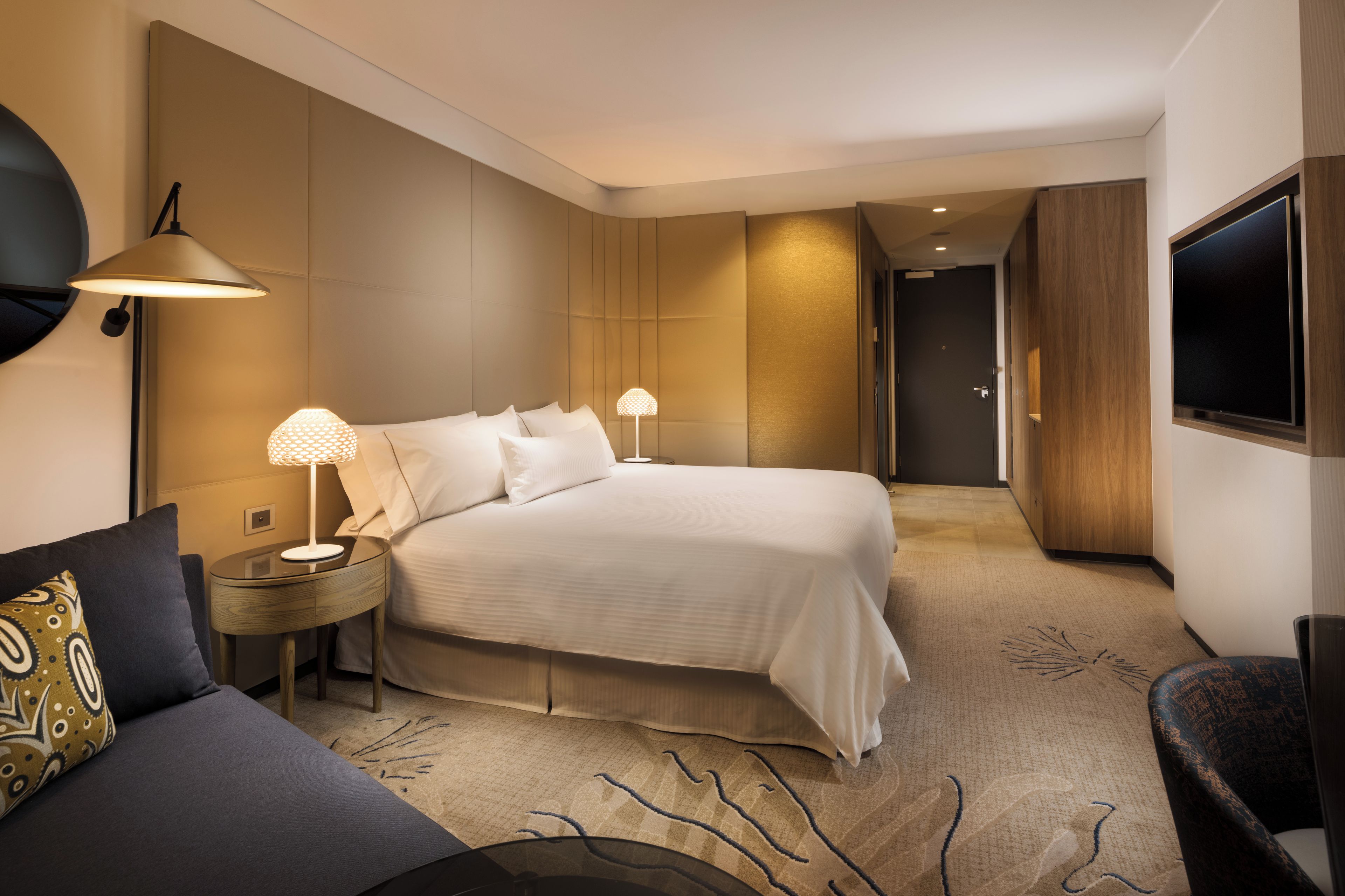 Una habitación de hotel moderna con una cama grande, dos lámparas de noche, un televisor de pantalla plana, un sofá y un armario de madera.