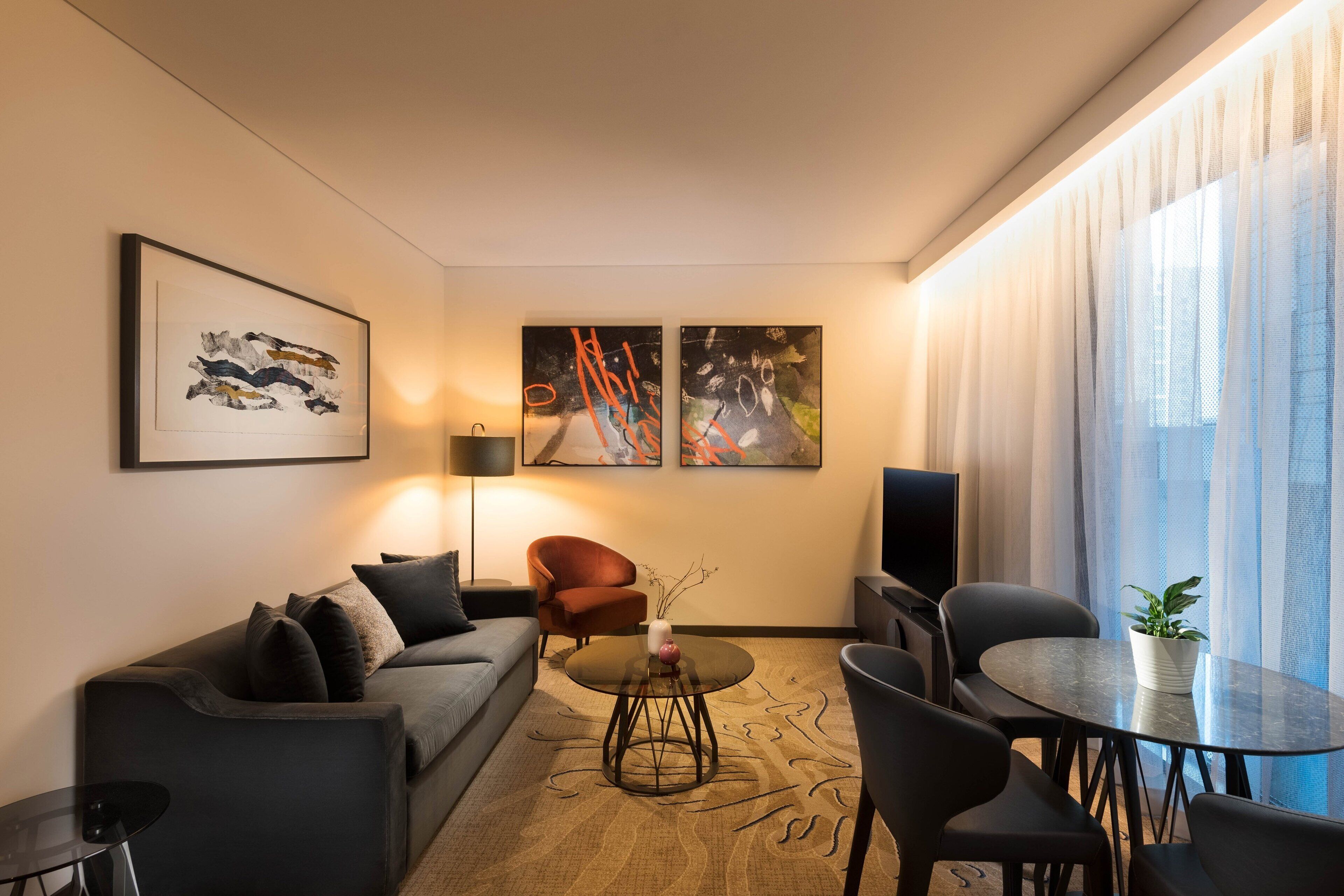Una sala de estar moderna con un sofá, sillones, una mesa de centro y un televisor. Hay cuadros abstractos en la pared y una ventana con cortinas translúcidas.