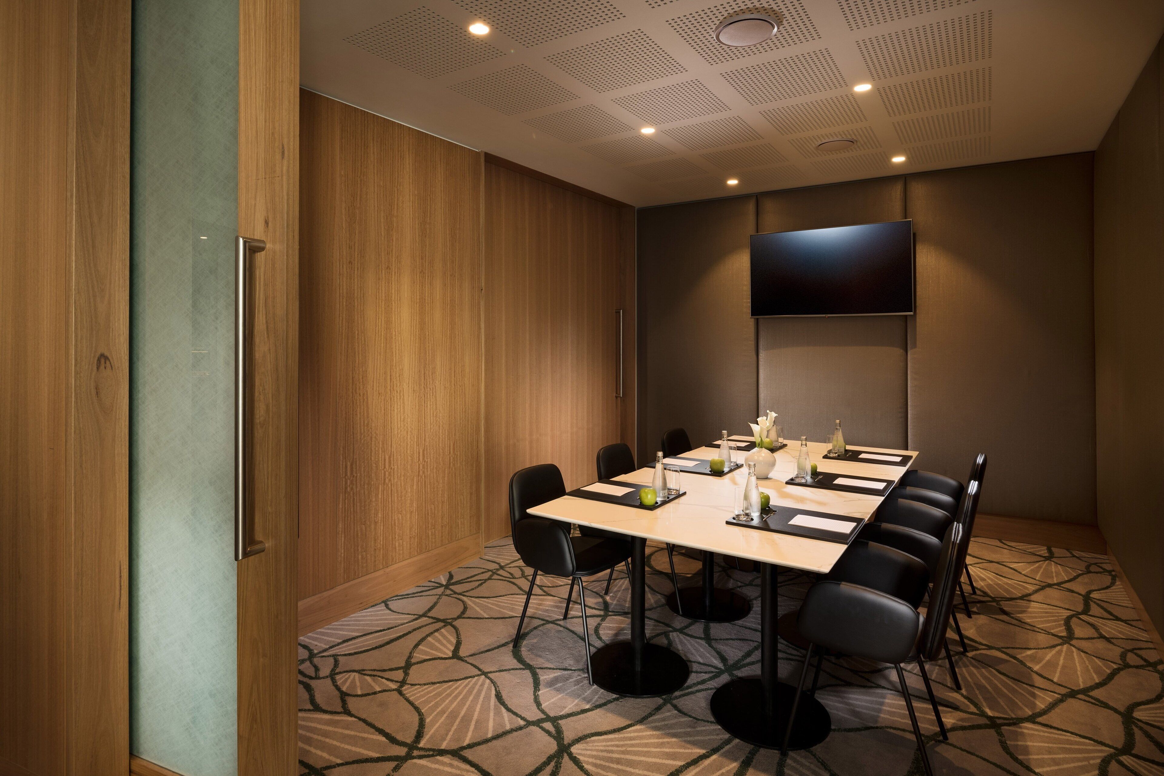 A conference room with a long table, black chairs, a flat-screen TV, and a patterned carpet.