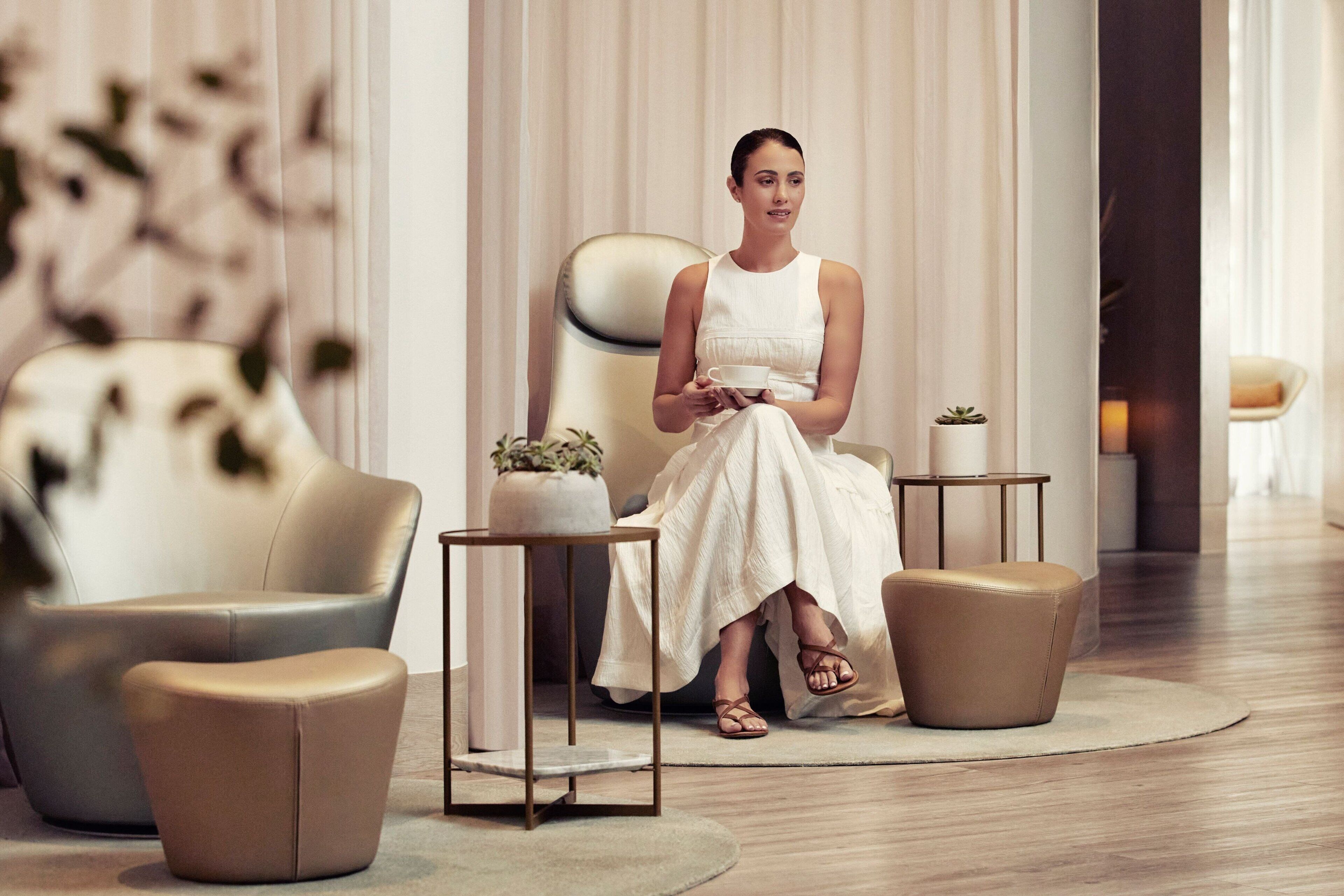 A woman seated on a sofa in a modern, well-lit room with a minimalist design, featuring a small table with a plant and a decorative item.