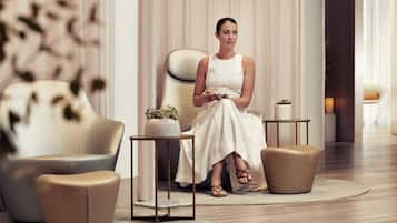 A woman seated on a sofa in a modern, well-lit room with a minimalist design, featuring a small table with a plant and a decorative item.
